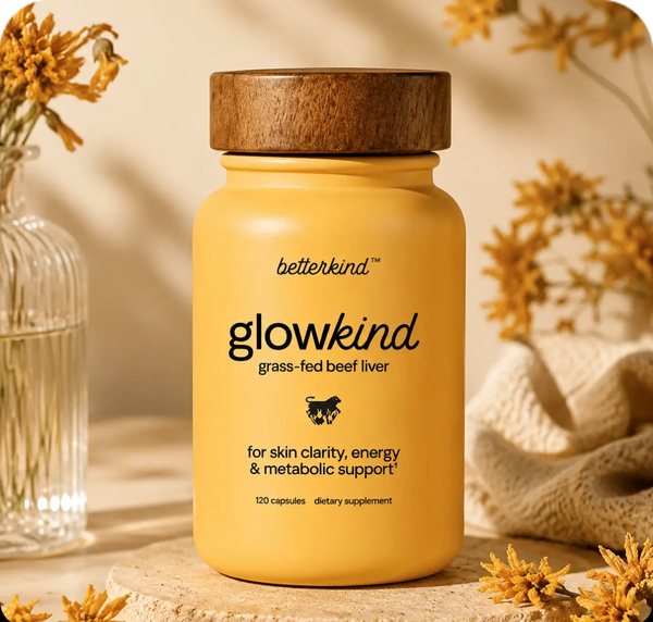 A yellow bottle of betterkind glowkind grass-fed beef liver dietary supplement with a wooden lid, styled on a stone surface with dried flowers and a woven cloth in a warm, natural setting.